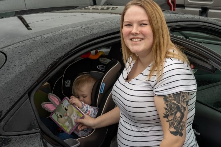 Woman standing outside car with a child in a carseat and a book